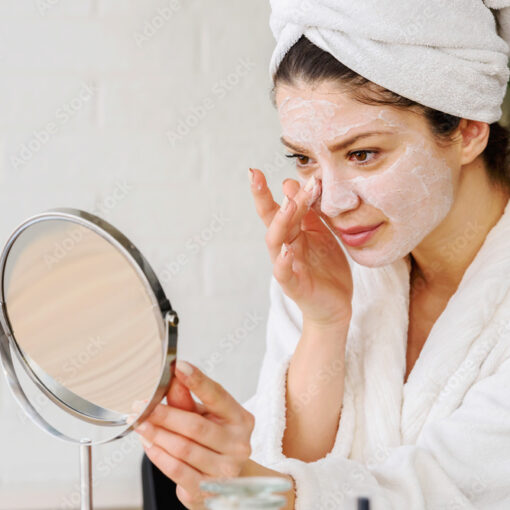 Eine Frau hat ein Handtuch auf dem Kopf und cremt ihr Gesicht ein während sie einen Spiegel hält