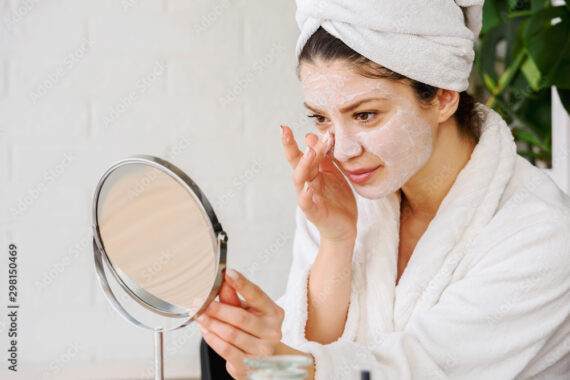 Eine Frau hat ein Handtuch auf dem Kopf und cremt ihr Gesicht ein während sie einen Spiegel hält